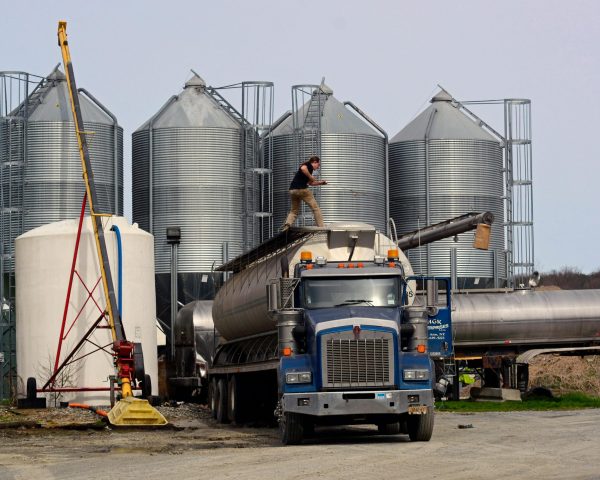 A worker loads a fuel truck in front of large industrial silos outdoors.