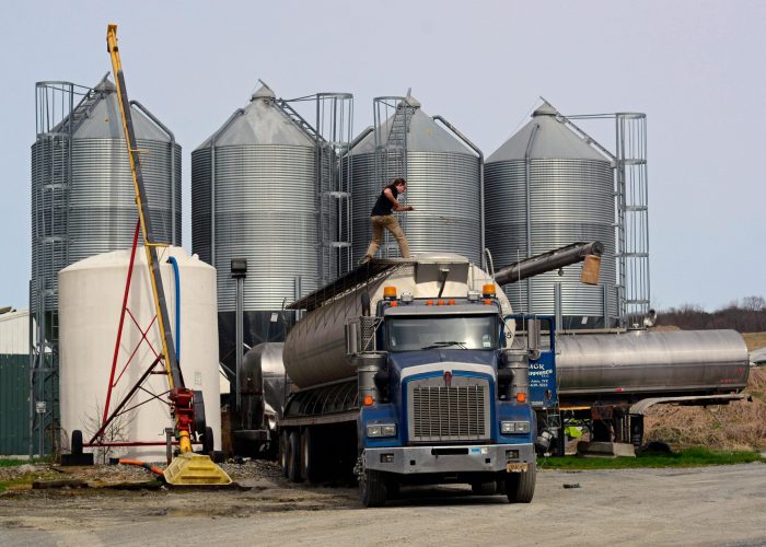 A worker loads a fuel truck in front of large industrial silos outdoors.