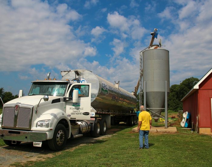 A fuel truck delivers to a silo on a farm under a blue sky.